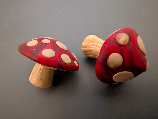 Two small red mushroom-shaped polymer clay models with beige spots on a gray background
